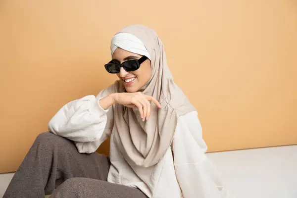 Mulher muçulmana feliz em hijab, óculos de sol e roupa casual na moda sentado e sorrindo no bege — Fotografia de Stock