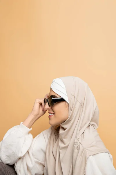 Side view of muslim woman in stylish sunglasses and hijab smiling on beige, modern individuality — Stock Photo