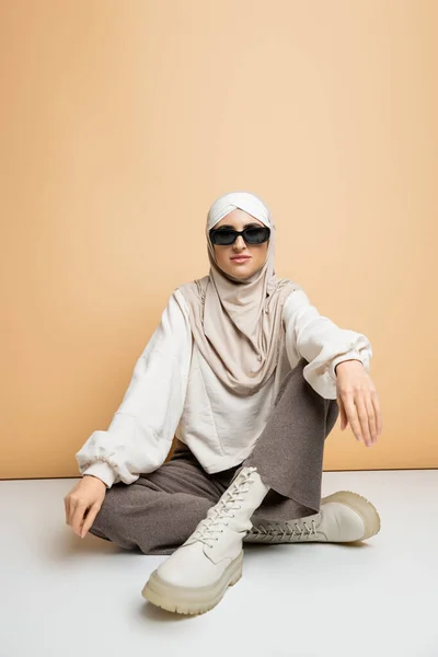 Donna musulmana indipendente in hijab, occhiali da sole e abbigliamento casual alla moda seduta su beige, lunghezza intera — Foto stock