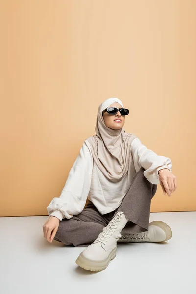 Modern style muslim woman in hijab, sunglasses and casual outfit sitting and smiling on beige — Stock Photo