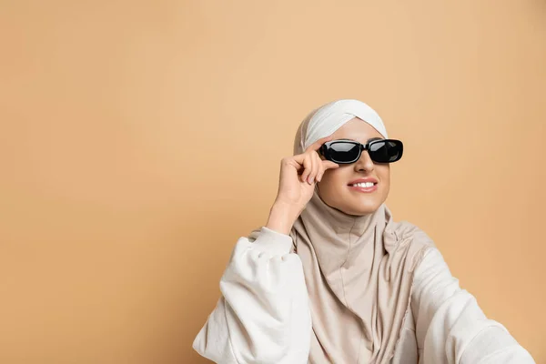 Mulher muçulmana sorridente em hijab e óculos de sol na moda olhando para longe em bege, auto-expressão moderna — Fotografia de Stock