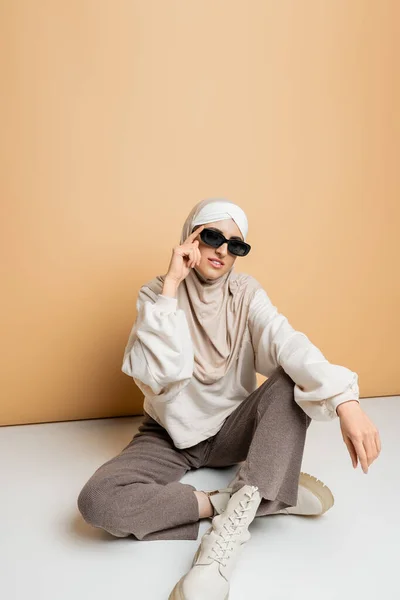 Comprimento total da mulher muçulmana em hijab e roupa casual na moda ajustando óculos de sol, sentado no bege — Fotografia de Stock