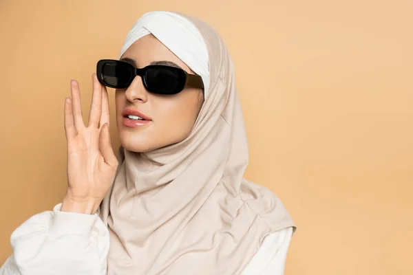 Portrait of modern muslim woman in hijab adjusting trendy sunglasses and looking away on beige — Stock Photo