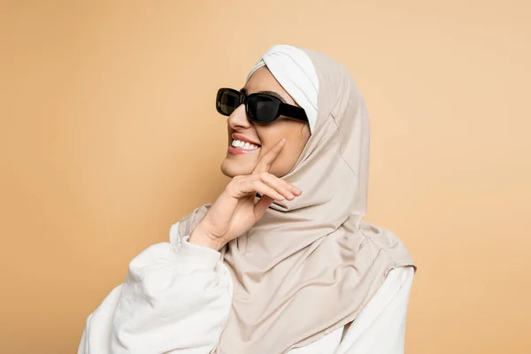 Mulher muçulmana sorridente em hijab e óculos de sol elegantes tocando rosto e olhando para longe em bege — Fotografia de Stock