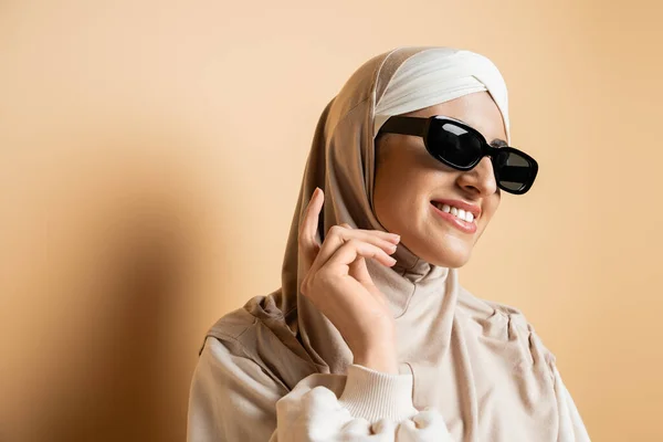 Moderne und lächelnde muslimische Frau in Hijab und trendiger Sonnenbrille, die wegschaut, während sie auf beige posiert — Stockfoto