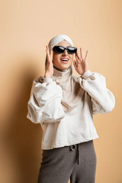 Donna musulmana stupita in hijab, felpa e abbigliamento casual alla moda che regola gli occhiali da sole sul beige — Foto stock
