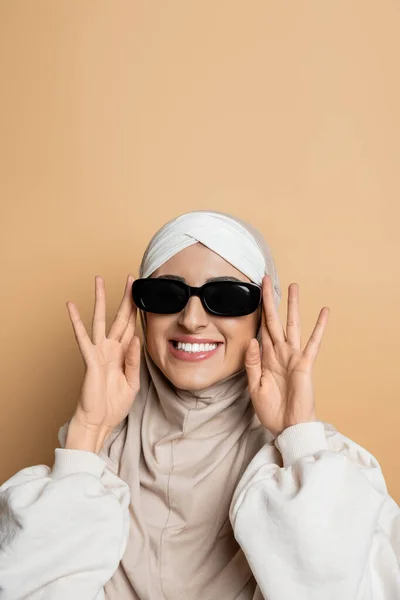 Mulher muçulmana alegre e elegante no hijab ajustando óculos de sol e sorrindo no bege — Fotografia de Stock