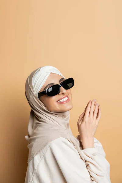 Mulher muçulmana satisfeito em hijab e óculos de sol na moda posando e sorrindo com as mãos orando em bege — Fotografia de Stock