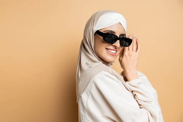 Fröhliche muslimische Frau im Hijab und stylischer Sonnenbrille lächelt in die Kamera, während sie auf Beige posiert — Stockfoto