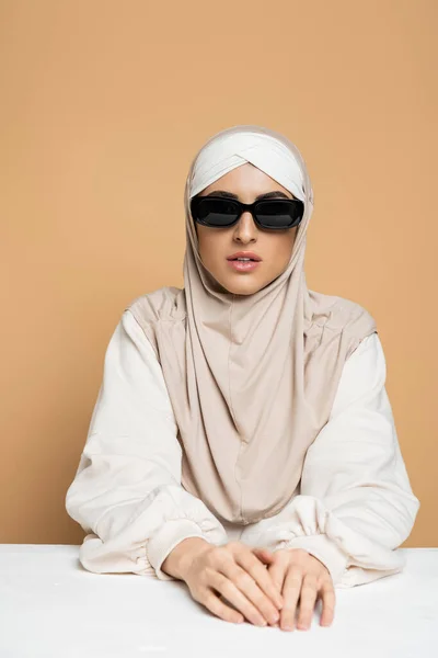 Confiante, mulher muçulmana na moda em hijab, óculos de sol e camisola sentada na mesa branca em bege — Fotografia de Stock