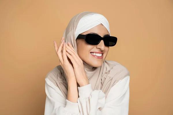 Joyous muslim woman in hijab, trendy sunglasses and white sweatshirt, with hands near face on beige — Stock Photo