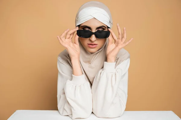 Trendige muslimische Frau in Hijab und Sweatshirt am weißen Tisch sitzend, Sonnenbrille abnehmend auf beige — Stockfoto