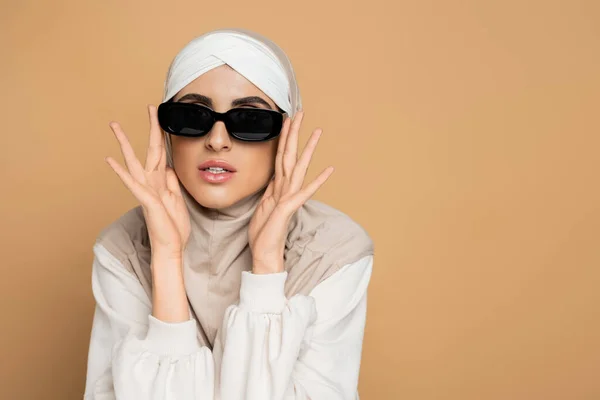 Mulher muçulmana moderna em hijab e camisola branca tocando óculos de sol elegantes enquanto posando em bege — Fotografia de Stock