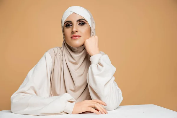 Mulher muçulmana na moda em hijab e camisola sentada à mesa, olhando para a mulher câmera no bege — Fotografia de Stock