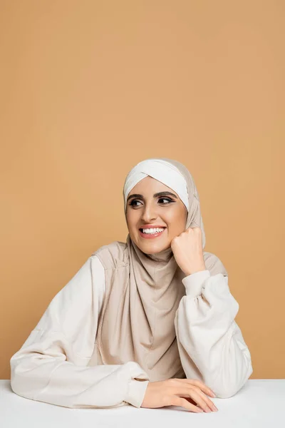 Carefree muslim woman in hijab and white sweatshirt sitting at white table and looking away on beige — Stock Photo