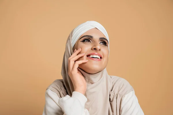 Mulher muçulmana animado em camisola branca e hijab segurando a mão perto do rosto e olhando para longe no bege — Fotografia de Stock