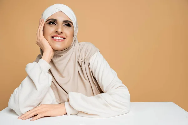 Mulher muçulmana feliz e sonhadora em camisola elegante e hijab olhando para a mesa branca no bege — Fotografia de Stock