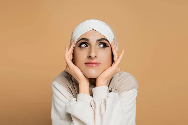 Retrato de mujer musulmana positiva y soñadora en hijab mirando hacia otro lado con las manos cerca de la cara en beige - foto de stock