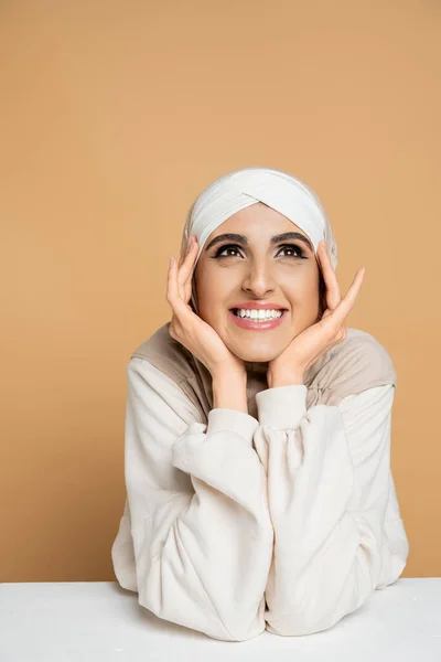 Happy and dreamy muslim woman in hijab holding hands near face and looking away at table on beige — Stock Photo