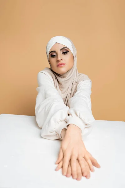 Pensativa mujer de Oriente Medio en hijab y sudadera sentado en la mesa blanca en beige, belleza musulmana - foto de stock
