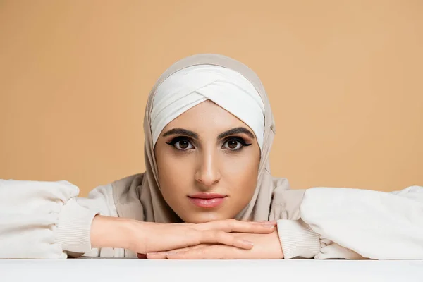 Mulher muçulmana atraente com maquiagem, sentado à mesa branca no hijab e olhando para a câmera no bege — Fotografia de Stock