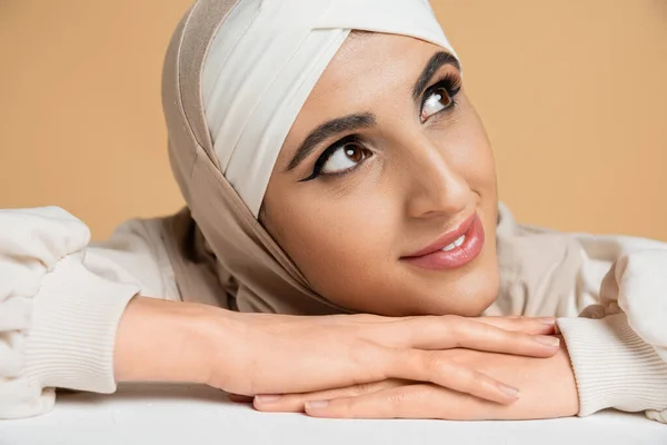 Primer plano retrato de mujer musulmana de ensueño con maquillaje, en hijab, sonriendo y mirando hacia otro lado en beige - foto de stock