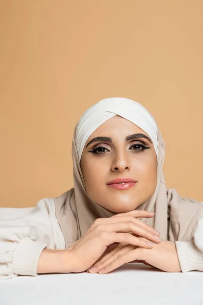 Mulher muçulmana positiva e elegante com maquiagem sentada à mesa branca no hijab e sorrindo para a câmera — Fotografia de Stock
