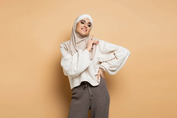 Donna musulmana allegra e moderna in hijab e abbigliamento casual alla moda in posa con mano sul fianco sul beige — Foto stock