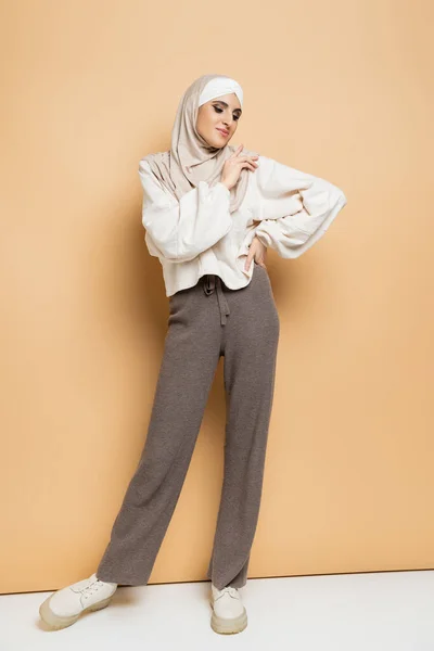 Fashionable muslim woman in hijab, white sweatshirt and grey pants posing with hand on hip on beige — Stock Photo