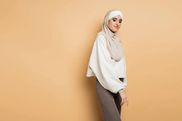 Charming muslim woman in hijab, white sweatshirt and grey pants standing on beige, personal style — Stock Photo