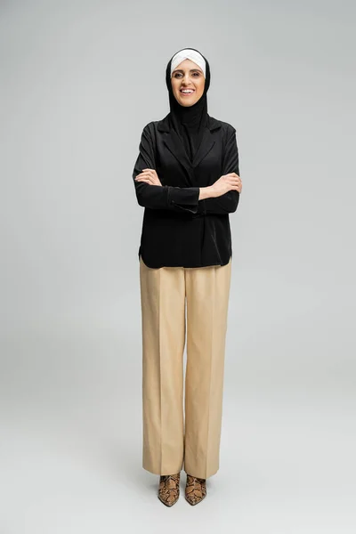 Happy muslim businesswoman in hijab and stylish outfit posing with folded arms on grey, full length — Stock Photo
