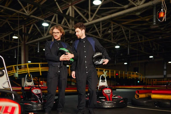 Due piloti di kart in piedi vicino alle auto da corsa e con in mano caschi, piloti maschi nel circuito di kart — Foto stock