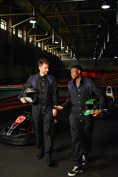 Coureurs de kart interracial marchant près des voitures de course et tenant des casques, les hommes à l'intérieur du circuit de kart — Photo de stock