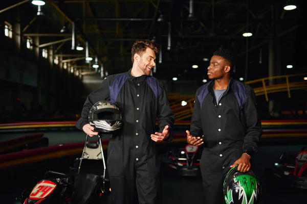 Heureux hommes multiculturels bavarder à l'intérieur du circuit de kart, coureurs de kart marchant et tenant des casques — Photo de stock