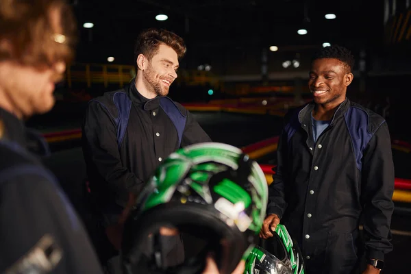 Homens multiculturais felizes conversando dentro do circuito de kart, três pilotos de kart segurando capacetes — Fotografia de Stock