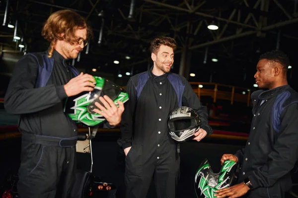 Hombres multiculturales positivos charlando dentro del circuito de kart, tres corredores de kart sosteniendo cascos - foto de stock