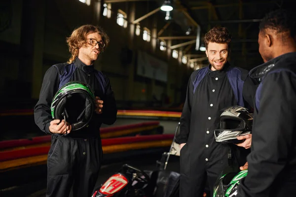 Positive interracial men chatting inside of kart circuit, three kart racers holding helmets — Stock Photo
