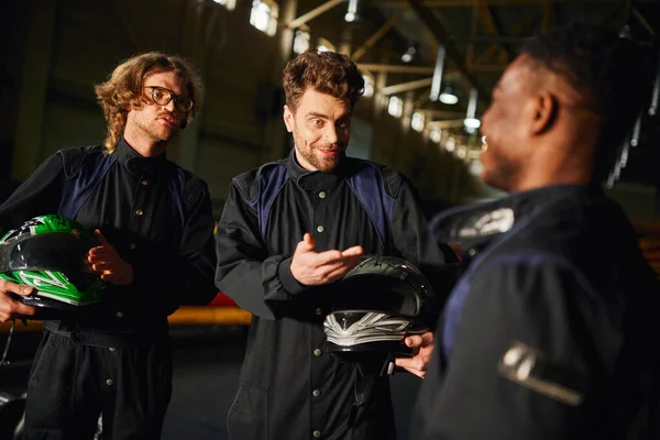 Conductores charlando con hombre afroamericano dentro del circuito de kart, tres corredores de kart con cascos - foto de stock