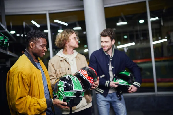 Felice uomo africano americano che tiene il casco vicino agli amici all'interno della pista da corsa al coperto, go-kart — Foto stock