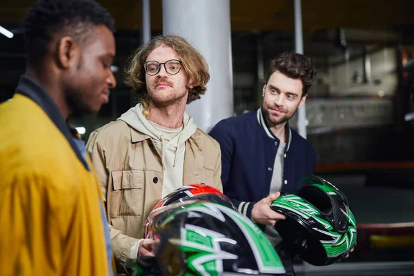 Hombres con cascos mirando a amigo afroamericano dentro de pista de carreras cubierta, karting - foto de stock