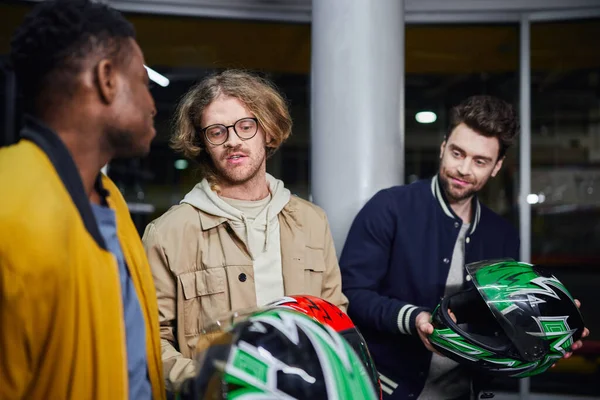 Uomini con caschi che chiacchierano con un amico afroamericano all'interno di una pista da corsa al coperto, karting — Foto stock