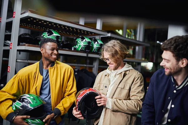 Fröhliche gemischtrassige männliche Freunde in Jacken mit Helmen, Indoor-Rennbahn, Kart-Konzept — Stockfoto