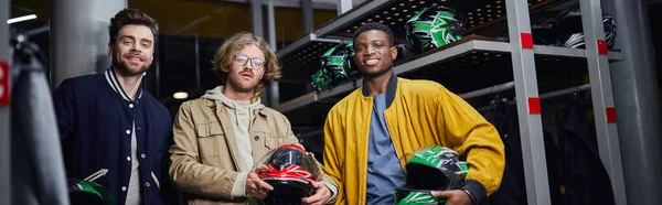 Happy multicultural friends holding helmets and looking at camera inside of racing track, banner — Stock Photo