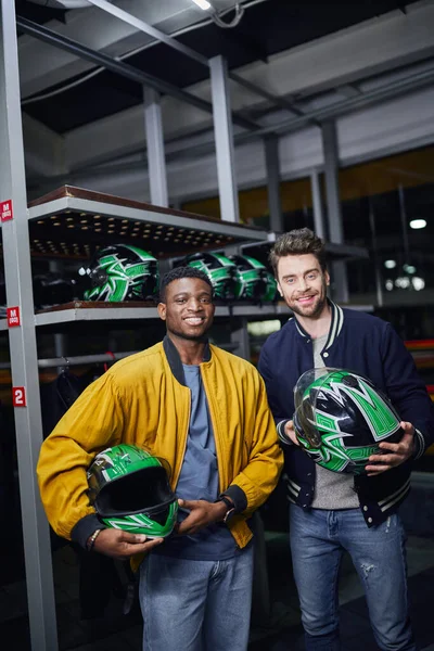 Dos amigos multiculturales sosteniendo cascos y mirando a la cámara dentro de la pista de karting, go-cart - foto de stock