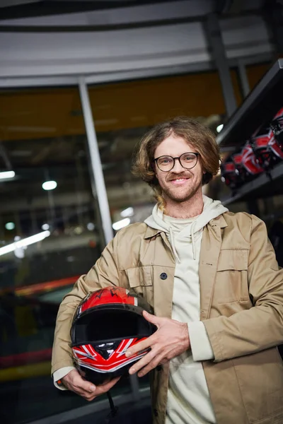 Felice uomo in occhiali che tiene il casco e guardando la fotocamera all'interno della pista di karting indoor, hobby — Foto stock