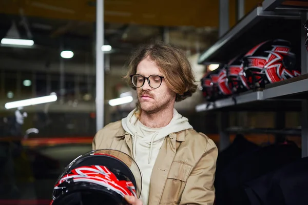 Homme en lunettes regardant casque à l'intérieur de piste de karting intérieur, sport automobile et passe-temps masculin — Photo de stock