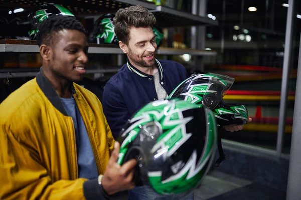Dos amigos multiculturales en chaquetas de bombardero mirando cascos dentro de la pista de karting, go-cart - foto de stock