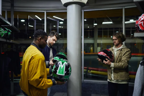 Tres amigos multiculturales en ropa casual eligiendo cascos para karting, concepto go-cart - foto de stock