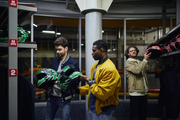 Drei gemischtrassige Freunde in lässiger Kleidung, die Helme für Kartfahren, Go-Cart-Konzept auswählen — Stockfoto