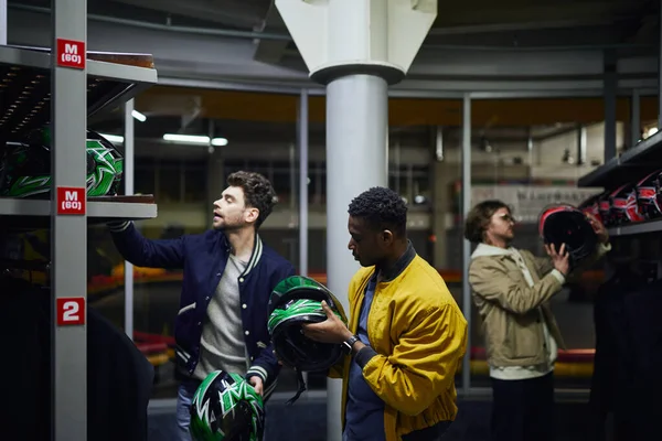 Trois hommes interraciaux en vêtements décontractés choisissant des casques pour le karting, concept go-cart, amis — Photo de stock
